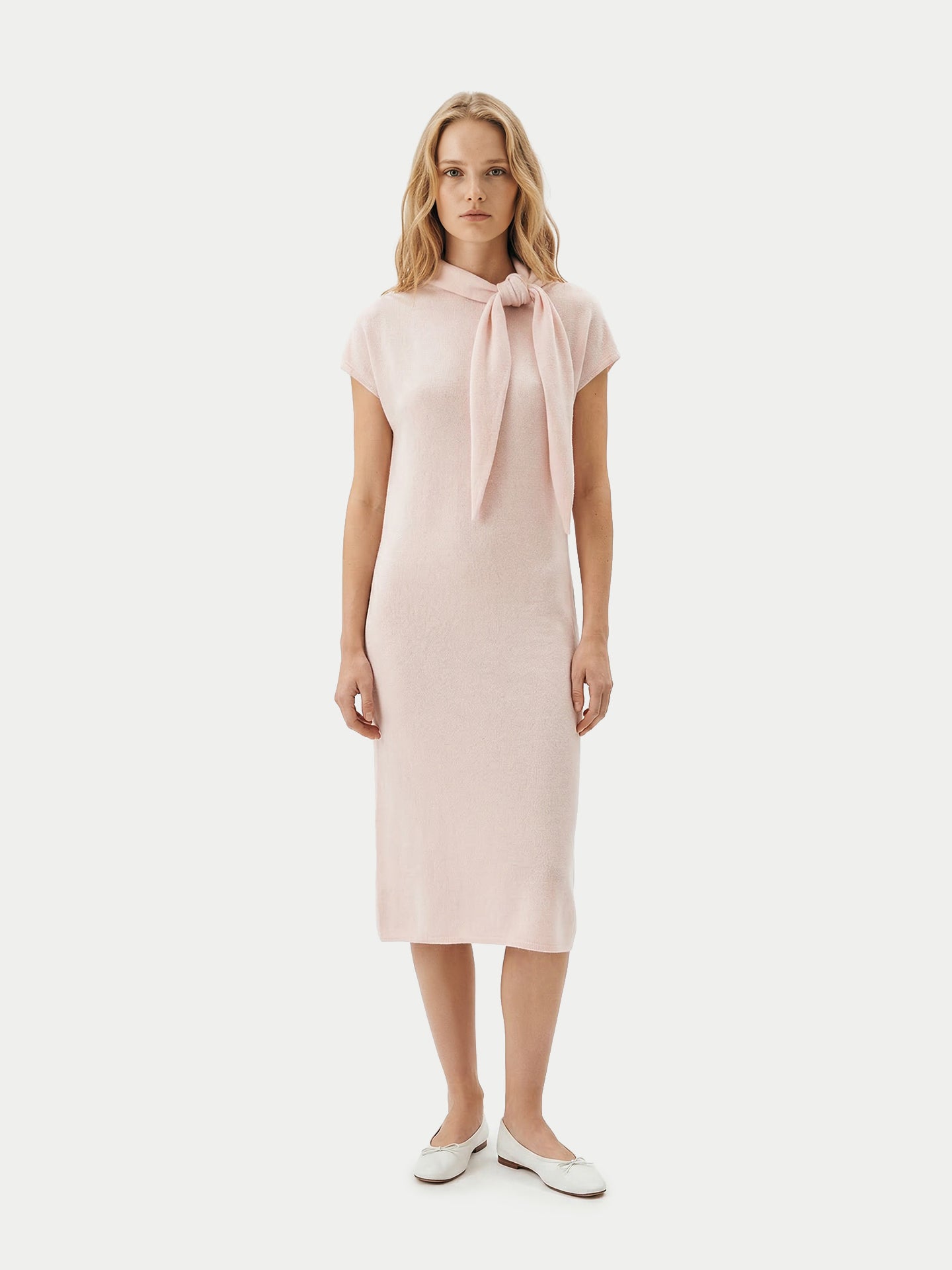 Vue de face d'une femme portant une robe rose arrivant sous le genou, avec un foulard noué et des ballerines blanches, sur un fond gris clair. #color_peach-blush