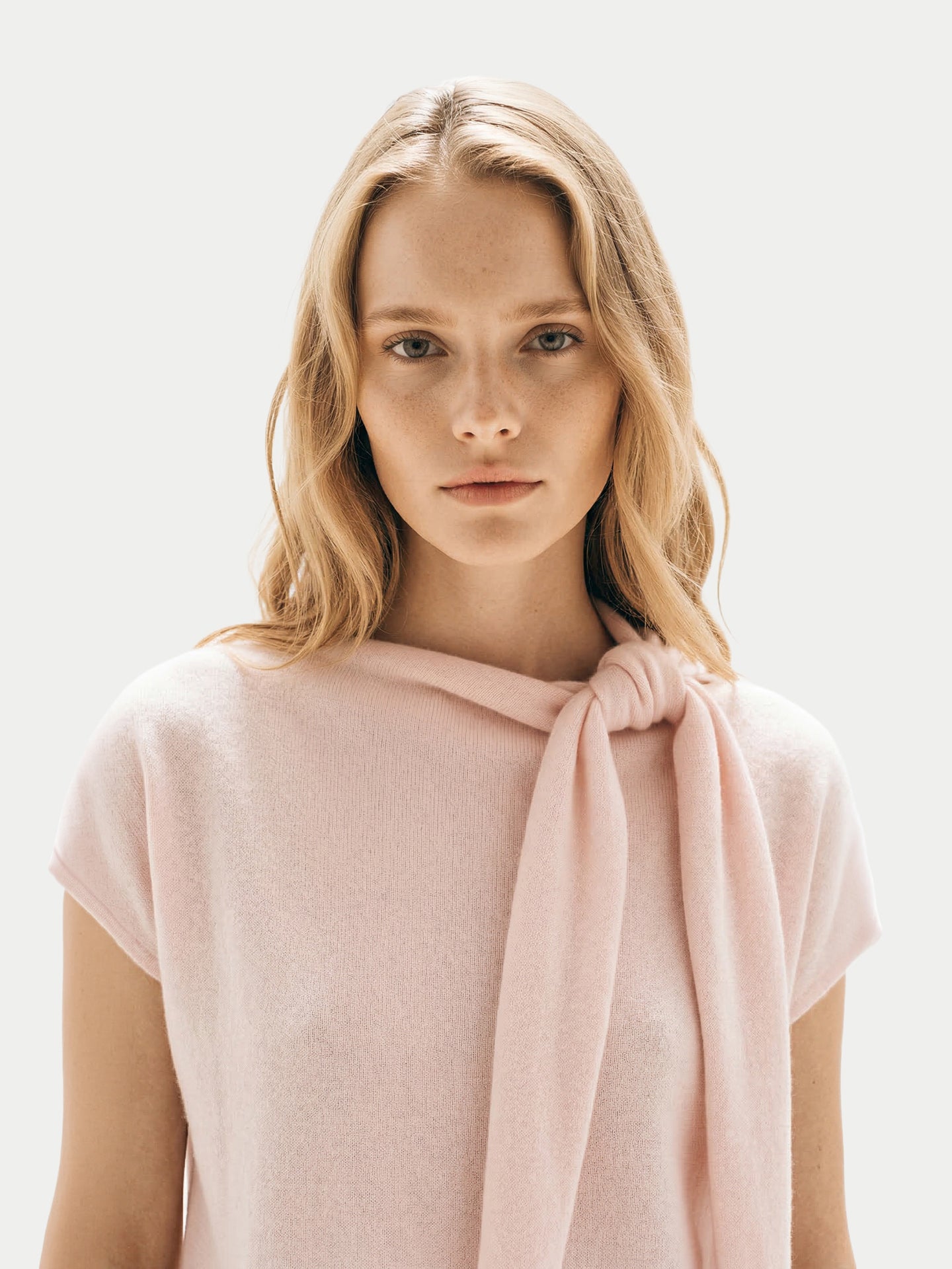 Gros plan de face d'une femme portant une robe rose arrivant sous le genou, avec un foulard noué et des ballerines blanches, sur un fond gris clair. #color_peach-blush