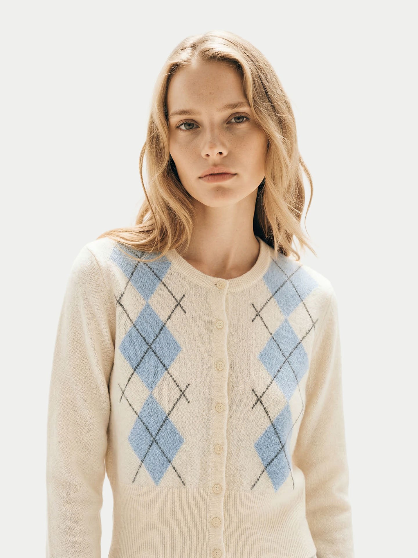 Gros plan de face d'une femme portant un cardigan blanc à col rond, à motifs losanges bleu clair, avec poignets et taille côtelés, sur fond gris clair. #color_blanc