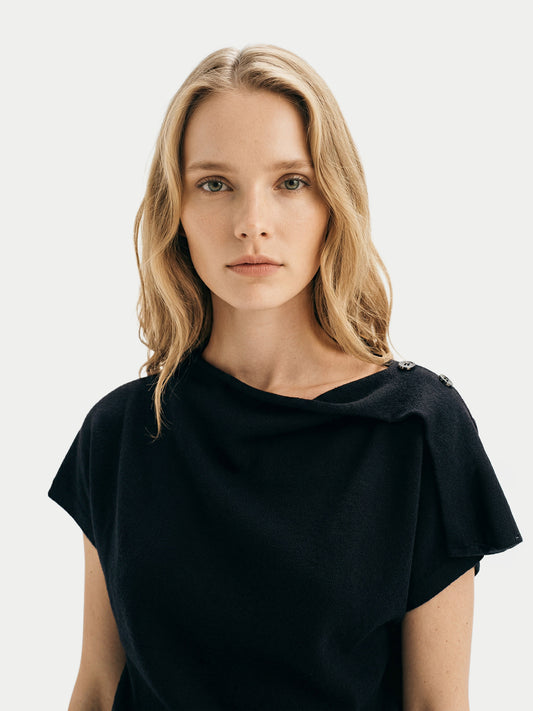 Gros plan de face d'une femme portant un haut en cachemire noir à manches courtes et col asymétrique sur un fond gris clair.#color_noir