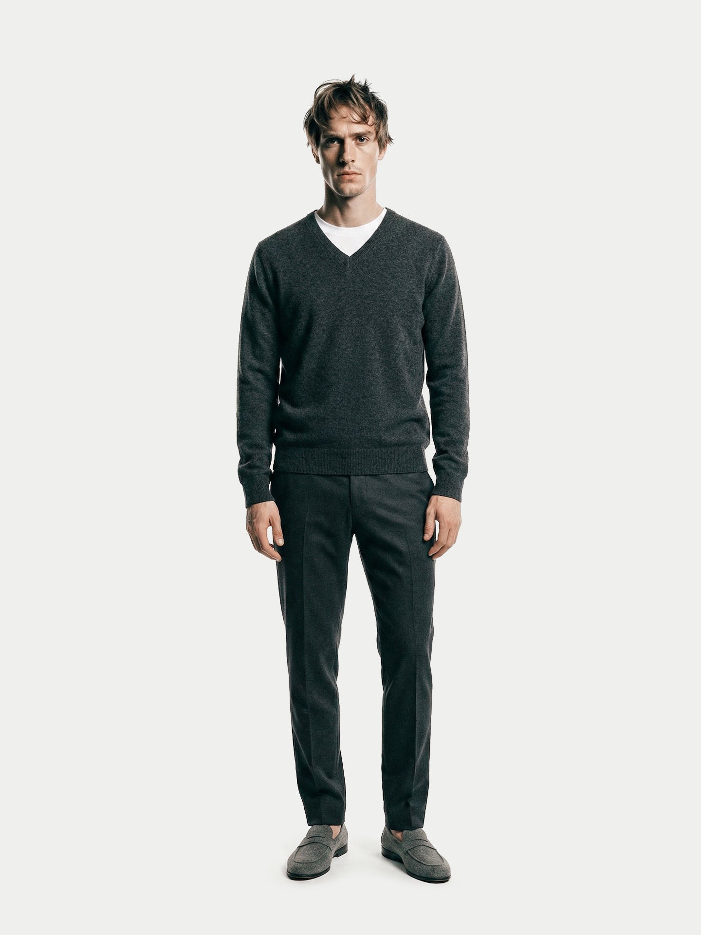 Vue de face d'un homme portant un pull gris foncé à col en V sur un t-shirt blanc et un pantalon noir, sur fond gris clair. #color_gris-taupe