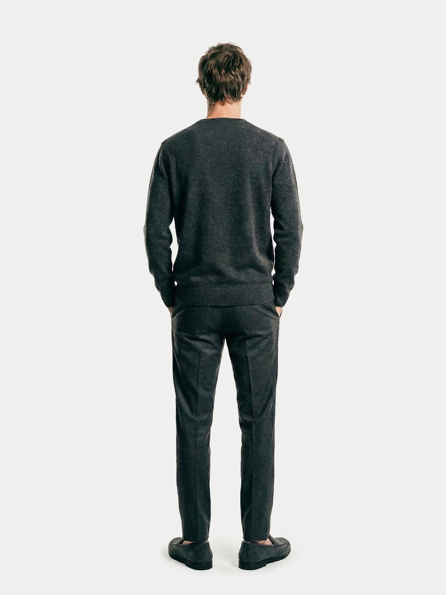 Vue de dos d'un homme portant un pull gris foncé sur un t-shirt blanc et un pantalon noir, sur fond gris clair. #color_gris-taupe