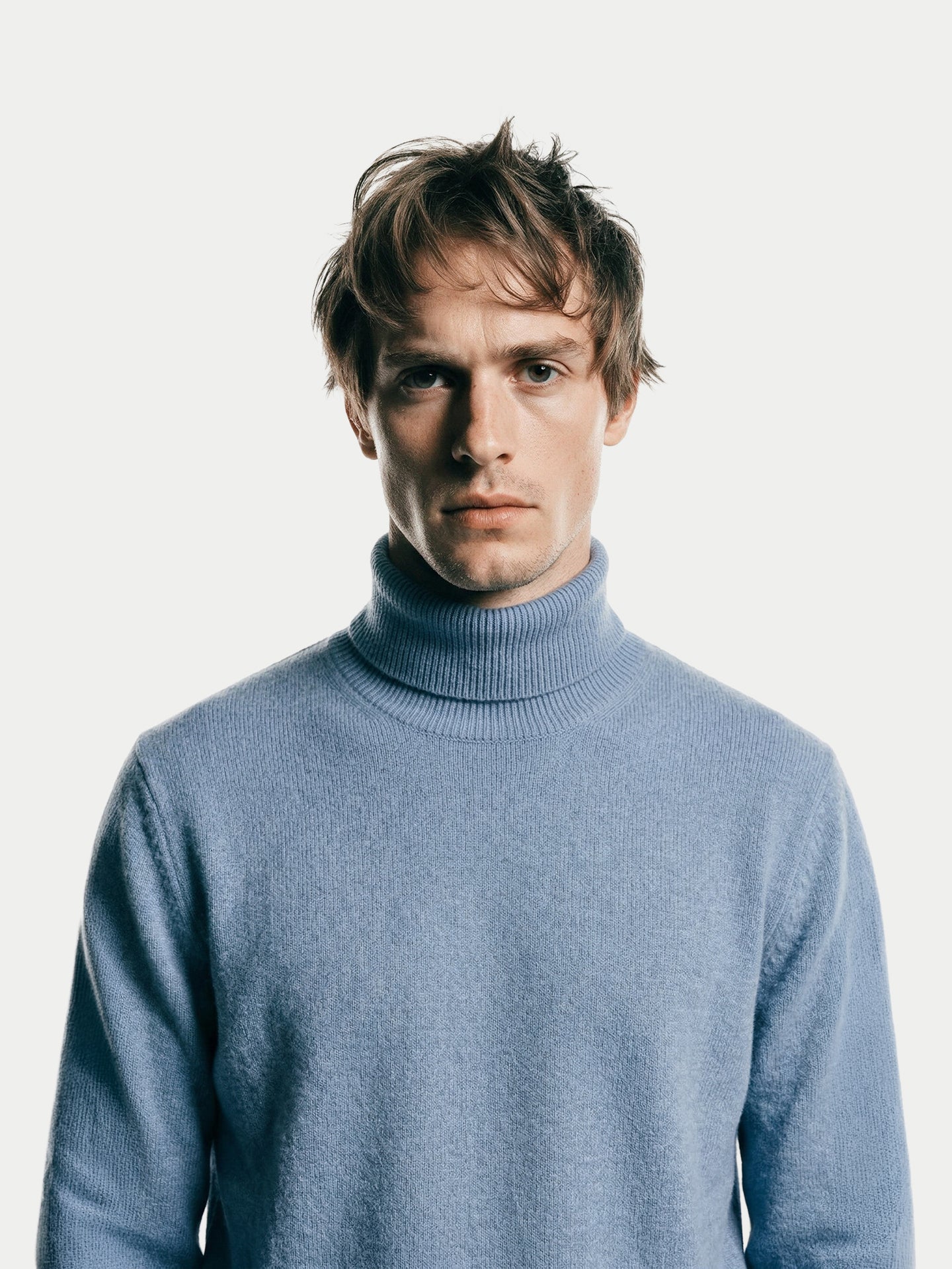 Gros plan de face d'un homme portant un pull à col roulé bleu clair sur fond gris clair. #color_bleu-clair