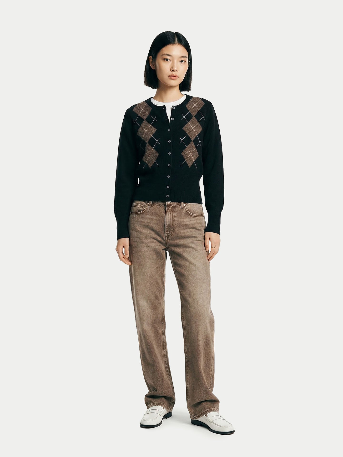 Vue de face d'une femme portant un cardigan noir à col rond, à motifs losanges marron, poignets et taille côtelés, sur un t-shirt blanc, un jean marron et des mocassins blancs, sur fond gris clair. #color_noir