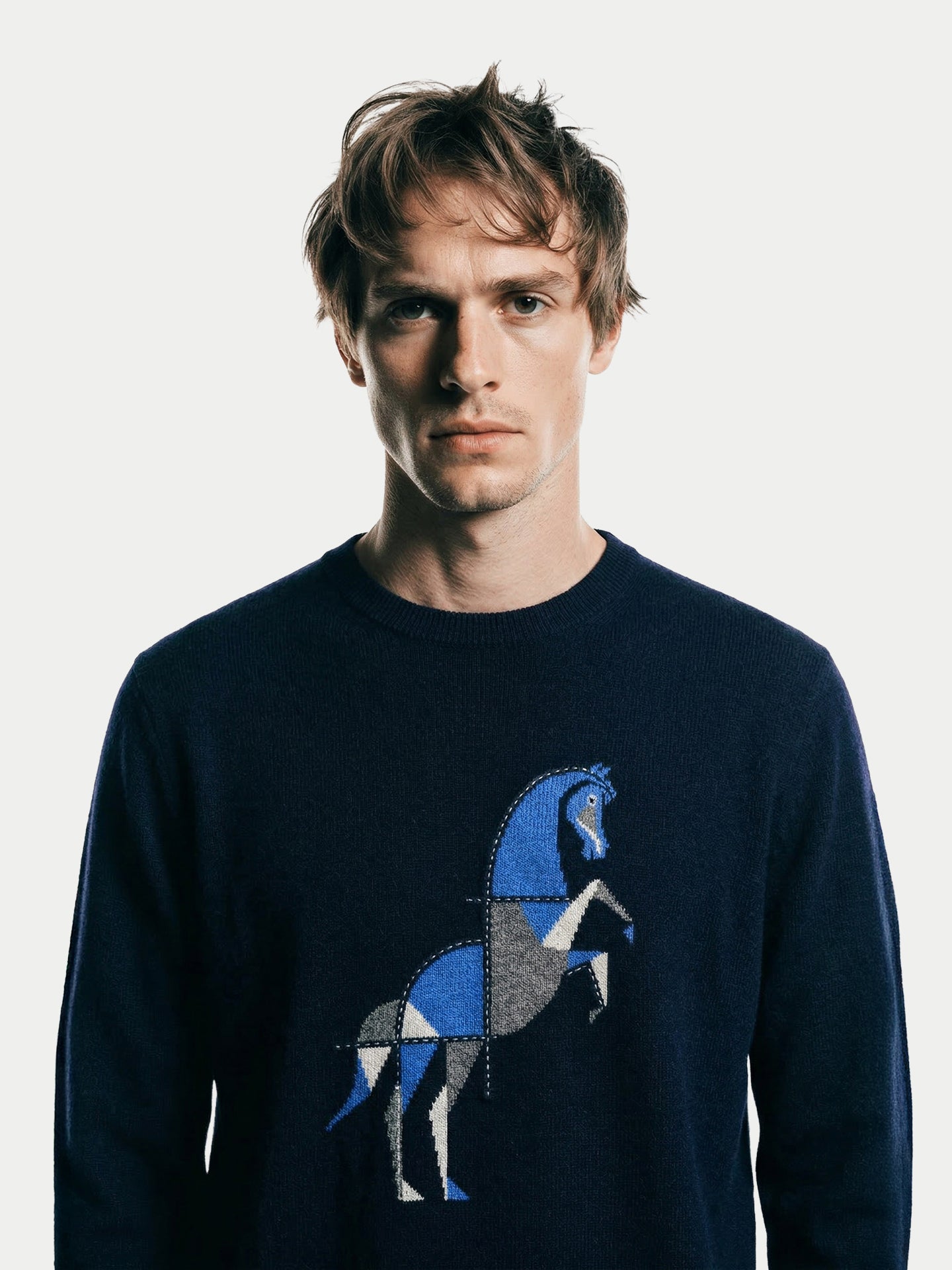 Gros plan de face d'un homme portant un pull bleu marine à col rond avec un motif de cheval sur fond gris clair #couleur_marine. #color_navy