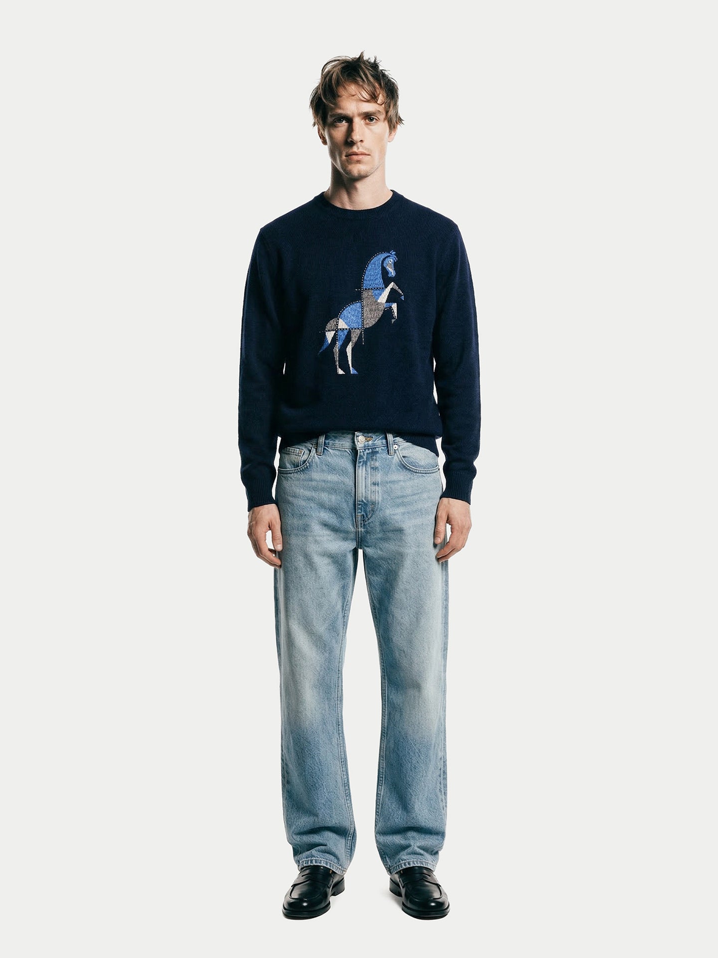 Vue de face d'un homme portant un pull bleu marine à col rond avec un motif de cheval et un jean bleu sur fond gris clair. #color_navy