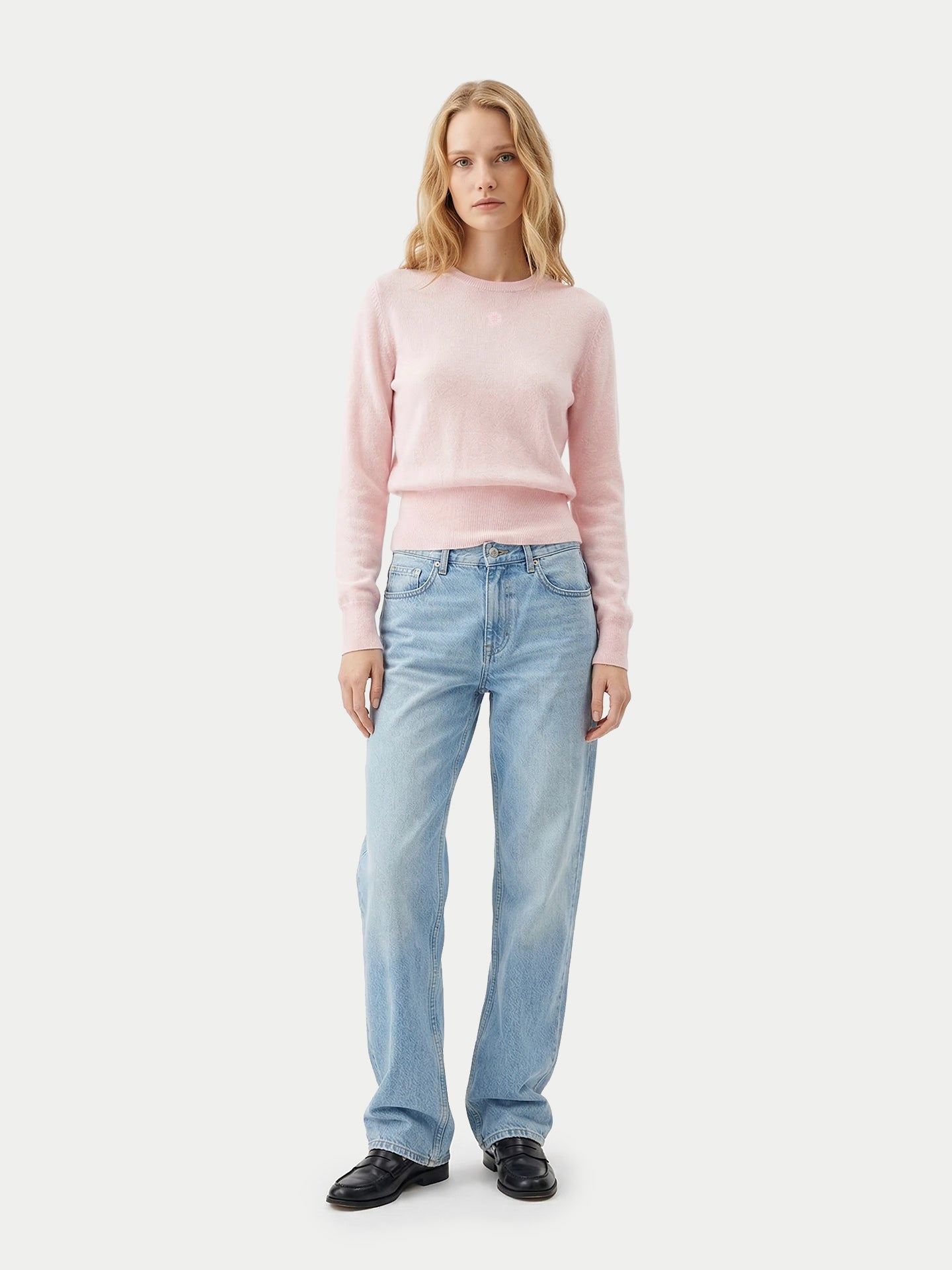 Vue de face d'une femme portant un pull rose clair à manches longues, poignets et taille côtelés, un jean bleu et des mocassins noirs sur fond gris clair. #color_peach-blush