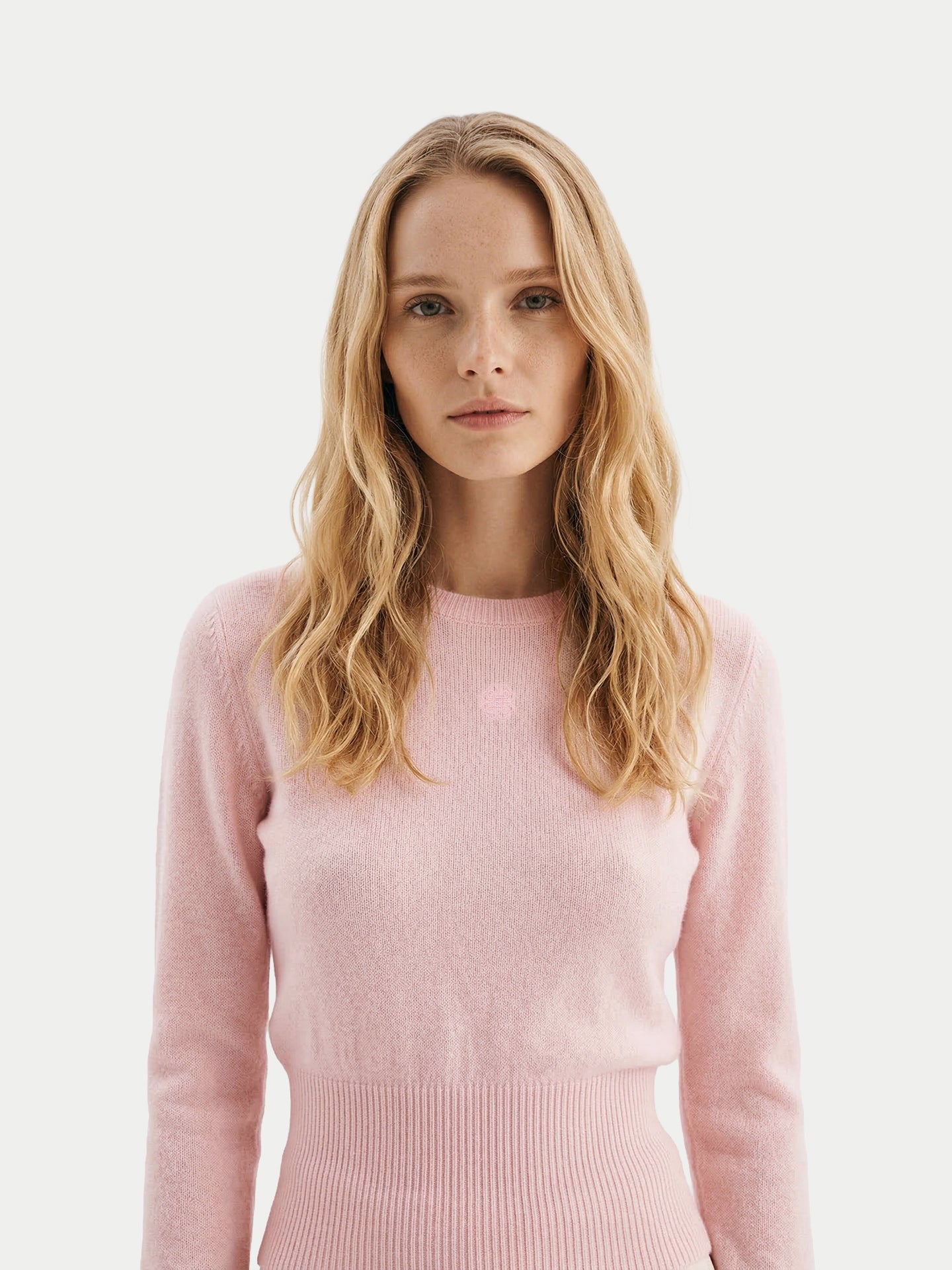 Gros plan de face d'une femme portant un pull rose clair à manches longues, avec poignets et taille côtelés, sur un fond gris clair. #color_peach-blush