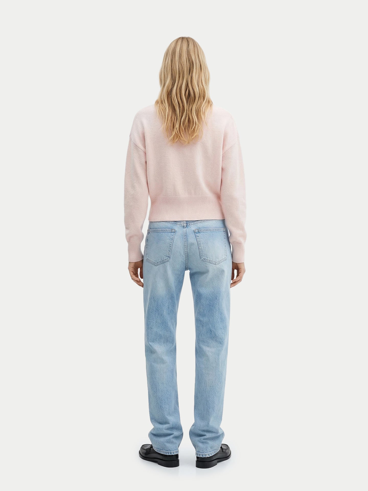 Vue de dos d'une femme portant un pull rose clair à manches longues, poignets et taille côtelés, un jean bleu et des chaussures noires sur un fond gris clair. #color_peach-blush