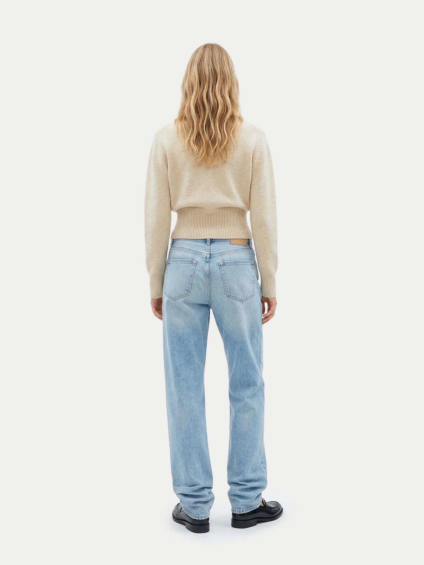 Vue de dos d'une femme portant un pull beige à manches longues, poignets et taille côtelés, un jean bleu et des chaussures noires sur un fond gris clair. #color_beige