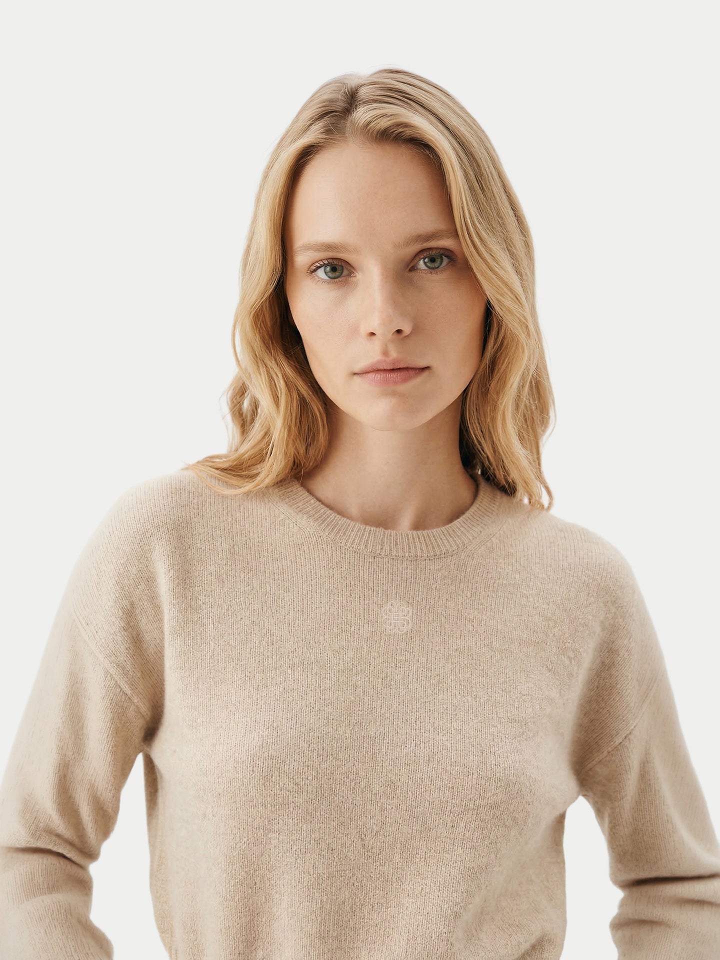 Gros plan de face d'un pull beige à manches longues avec un petit logo de marque en haut sur un fond gris clair. #color_beige