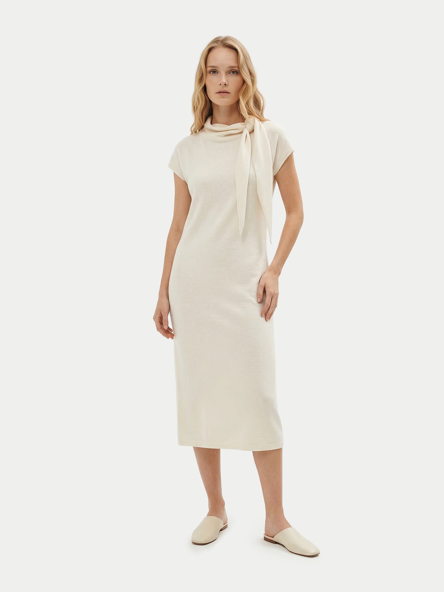 Vue de face d'une femme debout, vêtue d'une robe blanche à manches courtes arrivant sous le genou et portant un foulard noué, sur un fond gris clair. #color_blanc