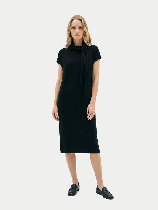 Vue de face d'une femme portant une robe noire à manches courtes arrivant sous le genou, avec un foulard noué, sur un fond gris clair. #color_noir