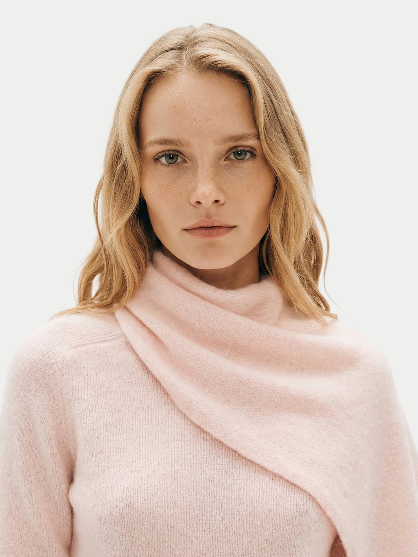 Gros plan de face d'une femme portant un pull rose avec une écharpe intégrée, sur fond gris clair. #color_peach-blush