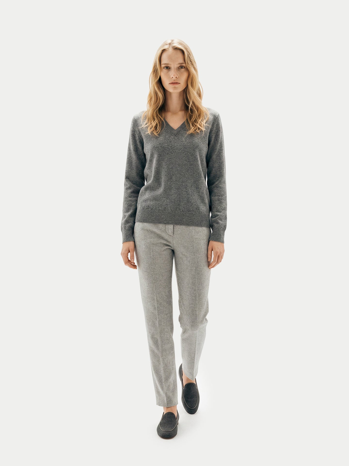  Vue de face d'une femme marchant, vêtue d'un pull gris foncé à col en V, d'un pantalon et de mocassins noirs, sur un fond gris clair. #color_gris-foncé