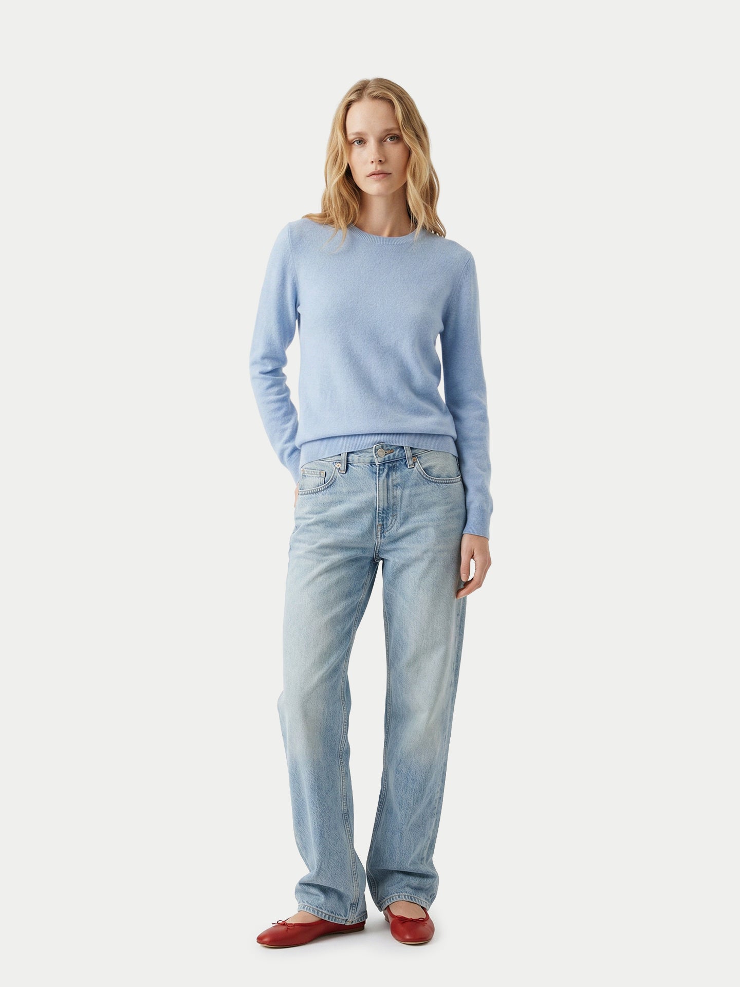 Vue de face d'une femme portant un pull ras du cou bleu clair, un jean bleu et des ballerines rouges sur un fond gris clair. #color_bleu-clair