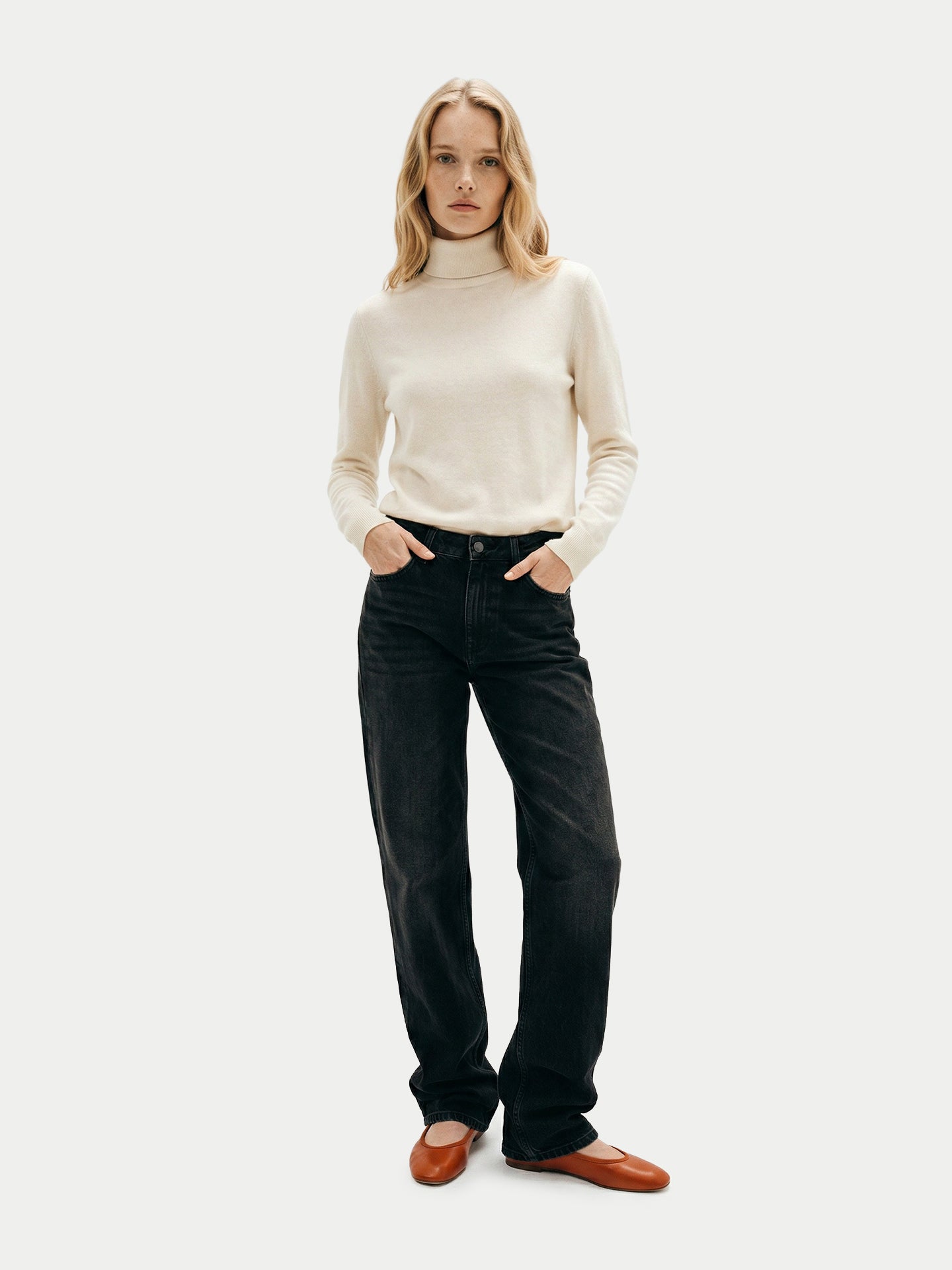 Vue de face d'une femme debout portant un pull à col roulé blanc, un jean noir et des ballerines rouges sur un fond gris clair. #color_blanc-cassé