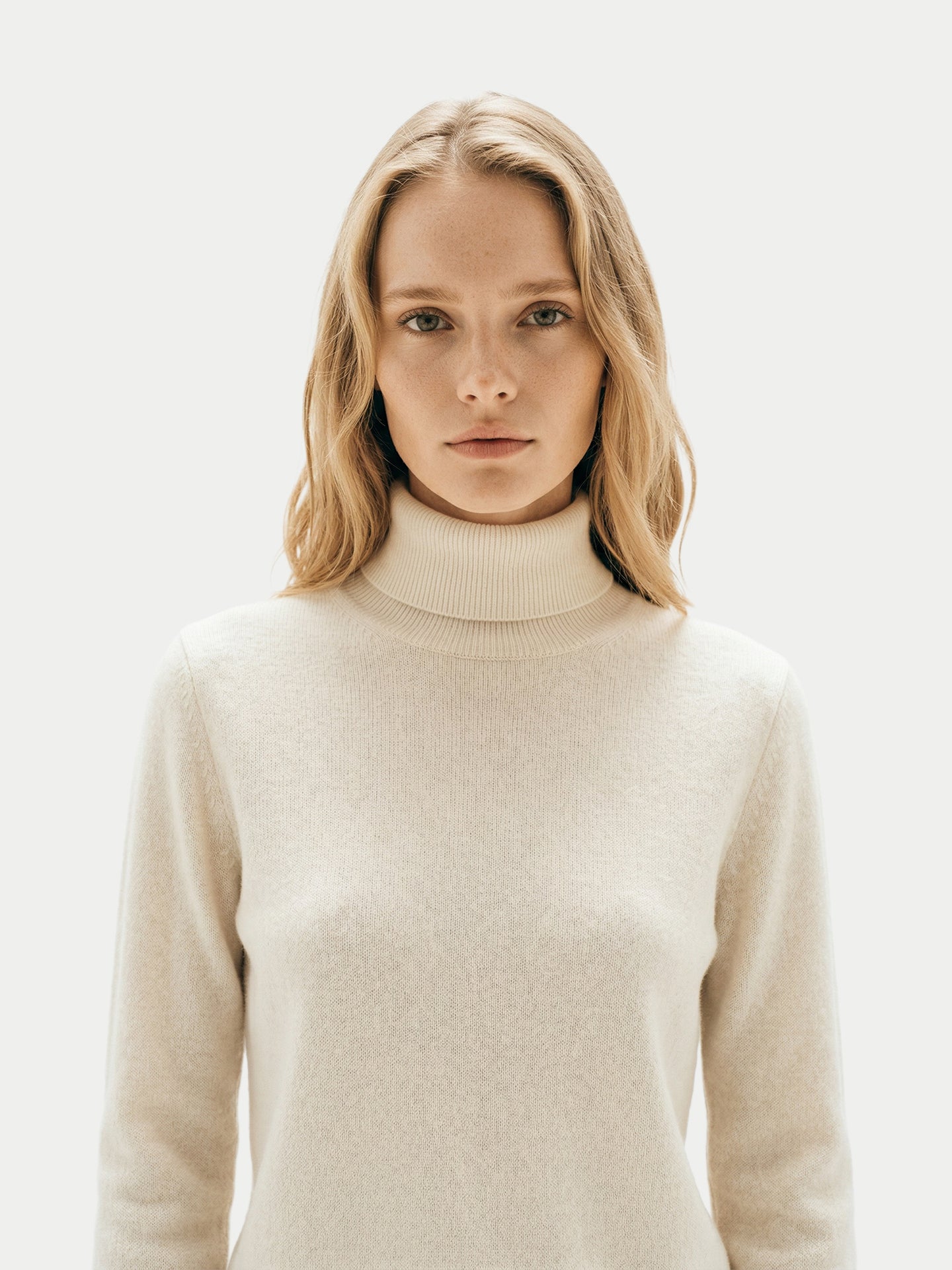 Gros plan de face d'une femme portant un pull à col roulé blanc sur fond gris clair. #color_blanc-cassé