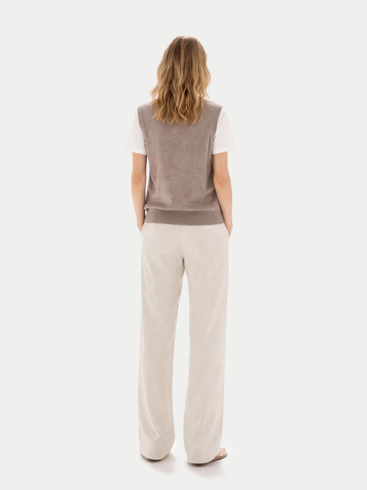 Vue de dos d'une femme portant un débardeur beige à col rond sur un t-shirt blanc et un pantalon blanc, sur fond gris clair. #color_taupe