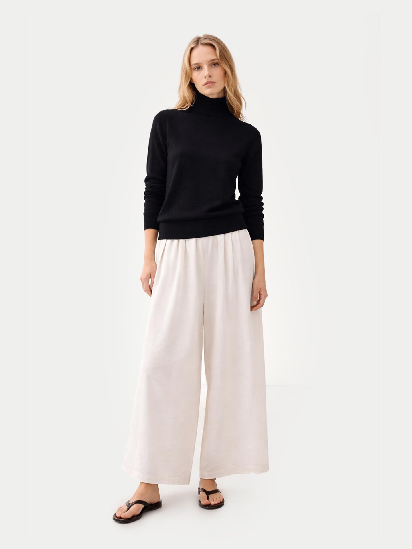 Vue de face d'une femme portant un pull à col roulé noir et un pantalon large sur fond gris clair. #color_noir