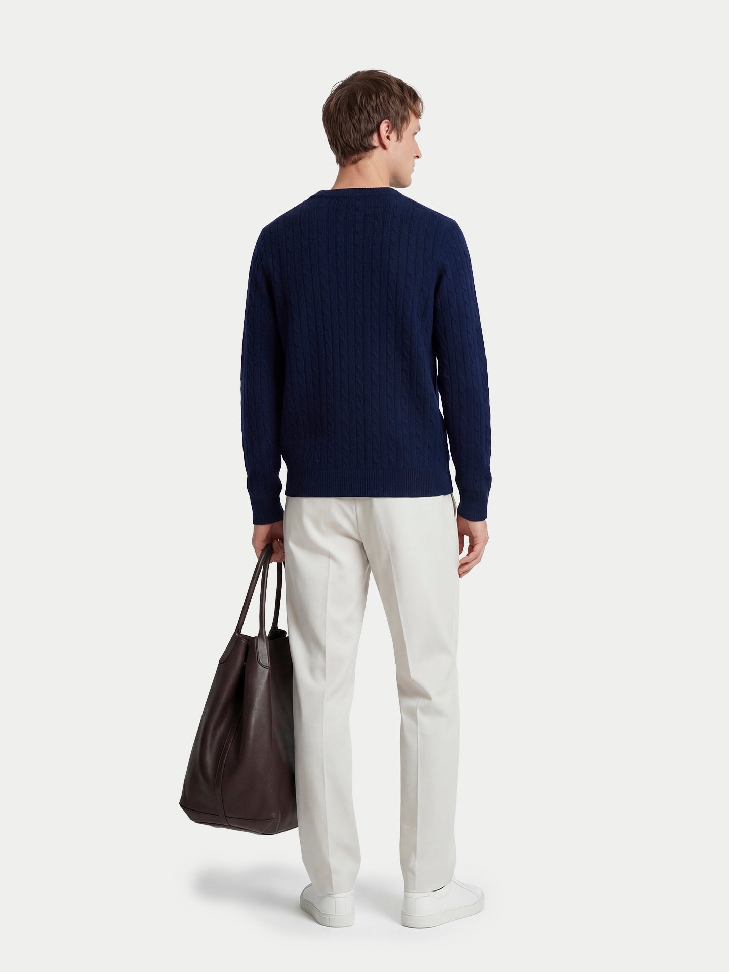 Vue de dos d'un homme debout, portant un pull en maille torsadée bleu marine, un pantalon blanc et tenant un sac à main, sur un fond gris clair. #color_navy