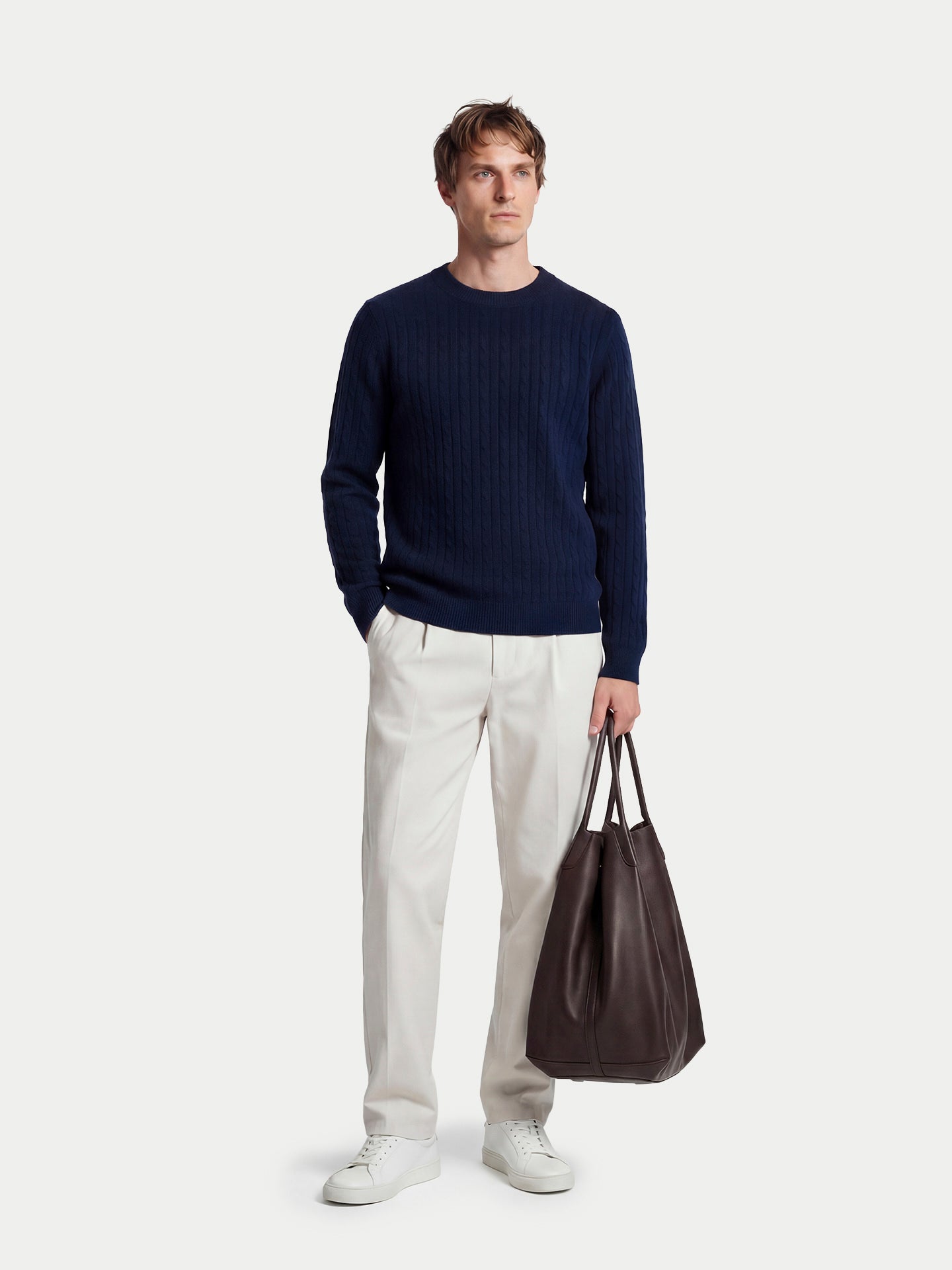 Vue de face d'un homme debout, portant un pull en maille torsadée bleu marine, un pantalon blanc et tenant un sac à main, sur un fond gris clair. #color_navy