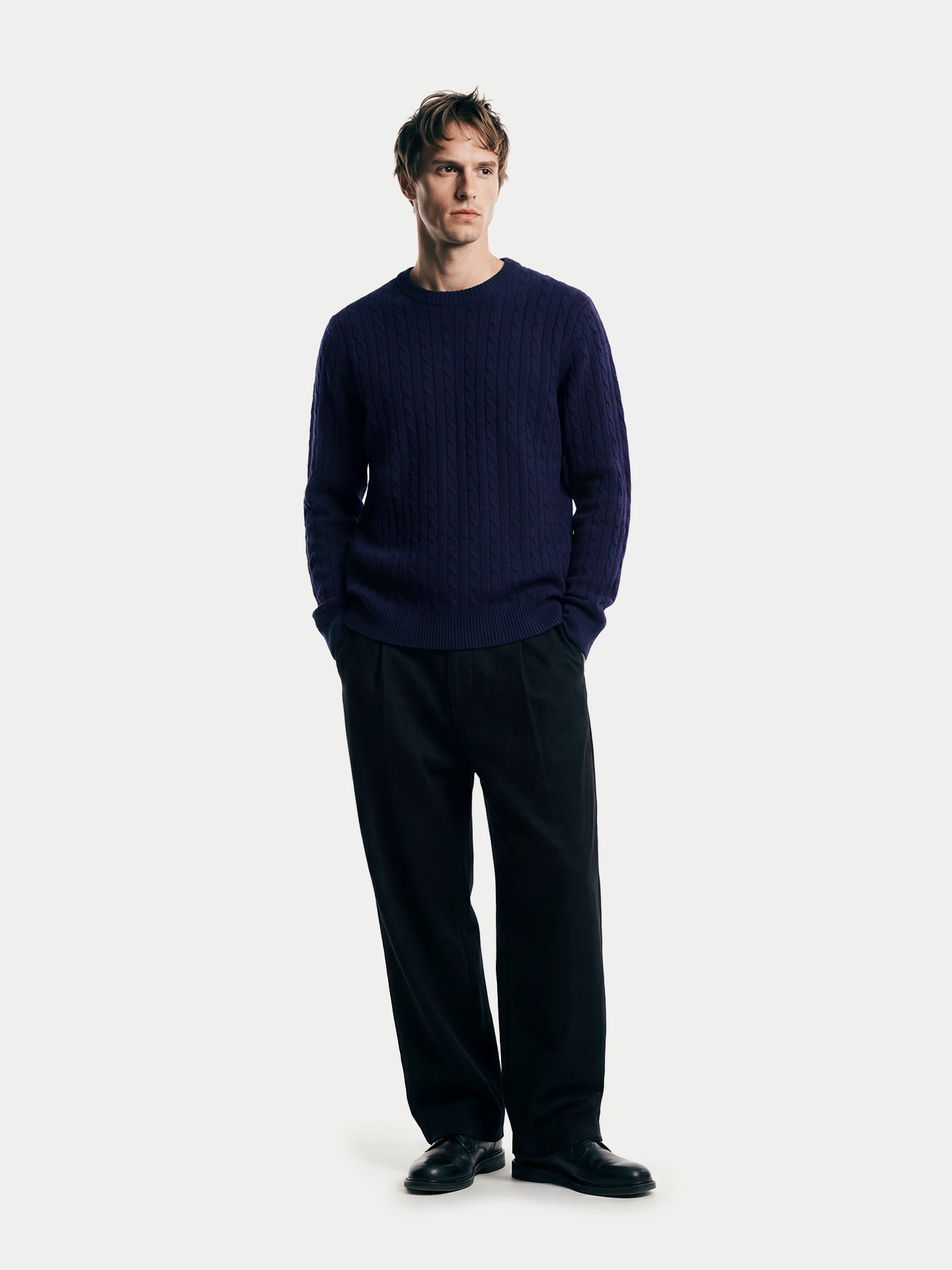 Vue de face d'un homme debout portant un pull en cachemire bleu marine à col rond et torsades, et un pantalon noir, sur fond gris clair. #color_navy