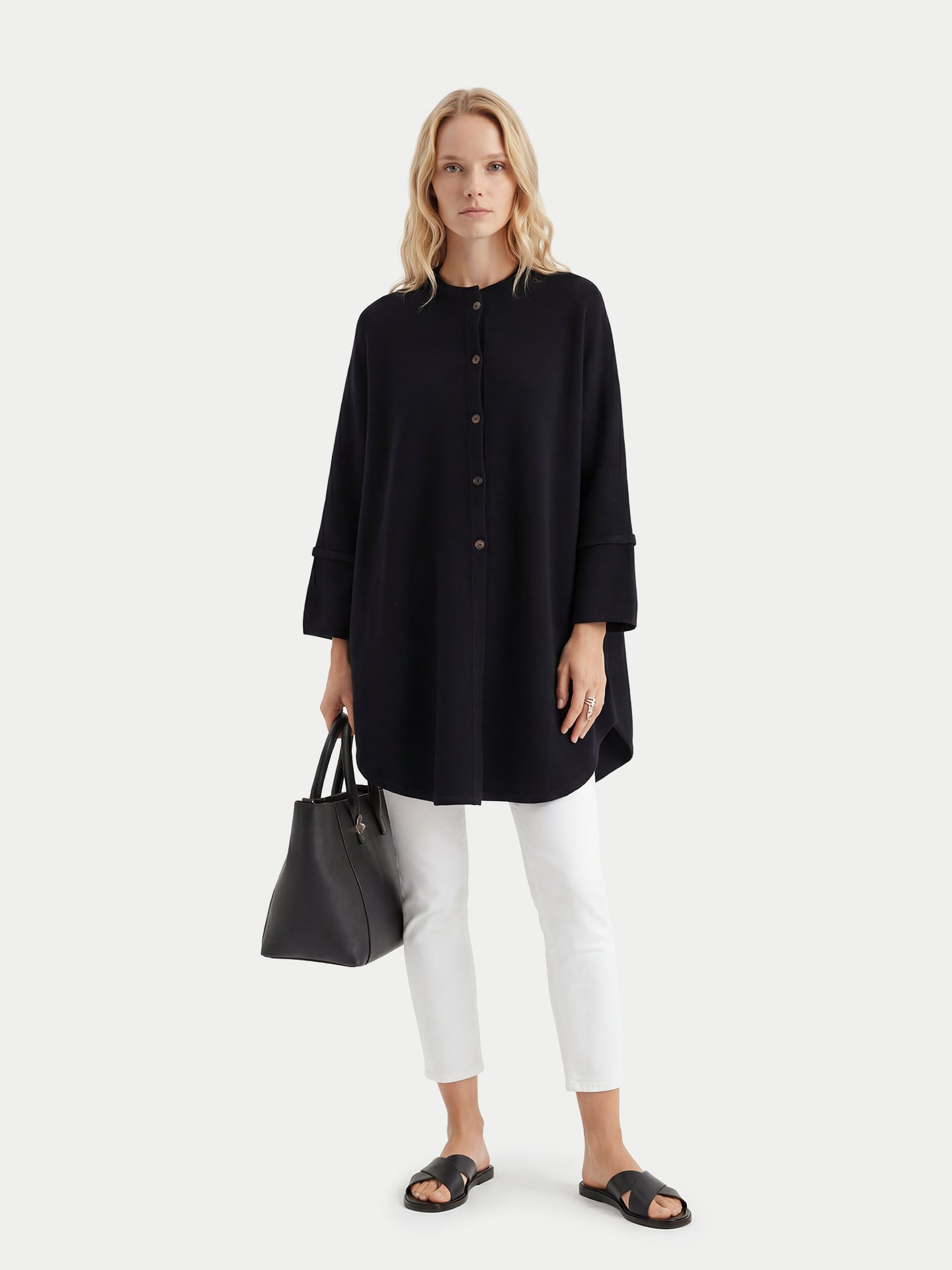 Femme portant un poncho noir à manches longues boutonné et un pantalon blanc, tenant un sac à main noir sur fond blanc. #color_noir