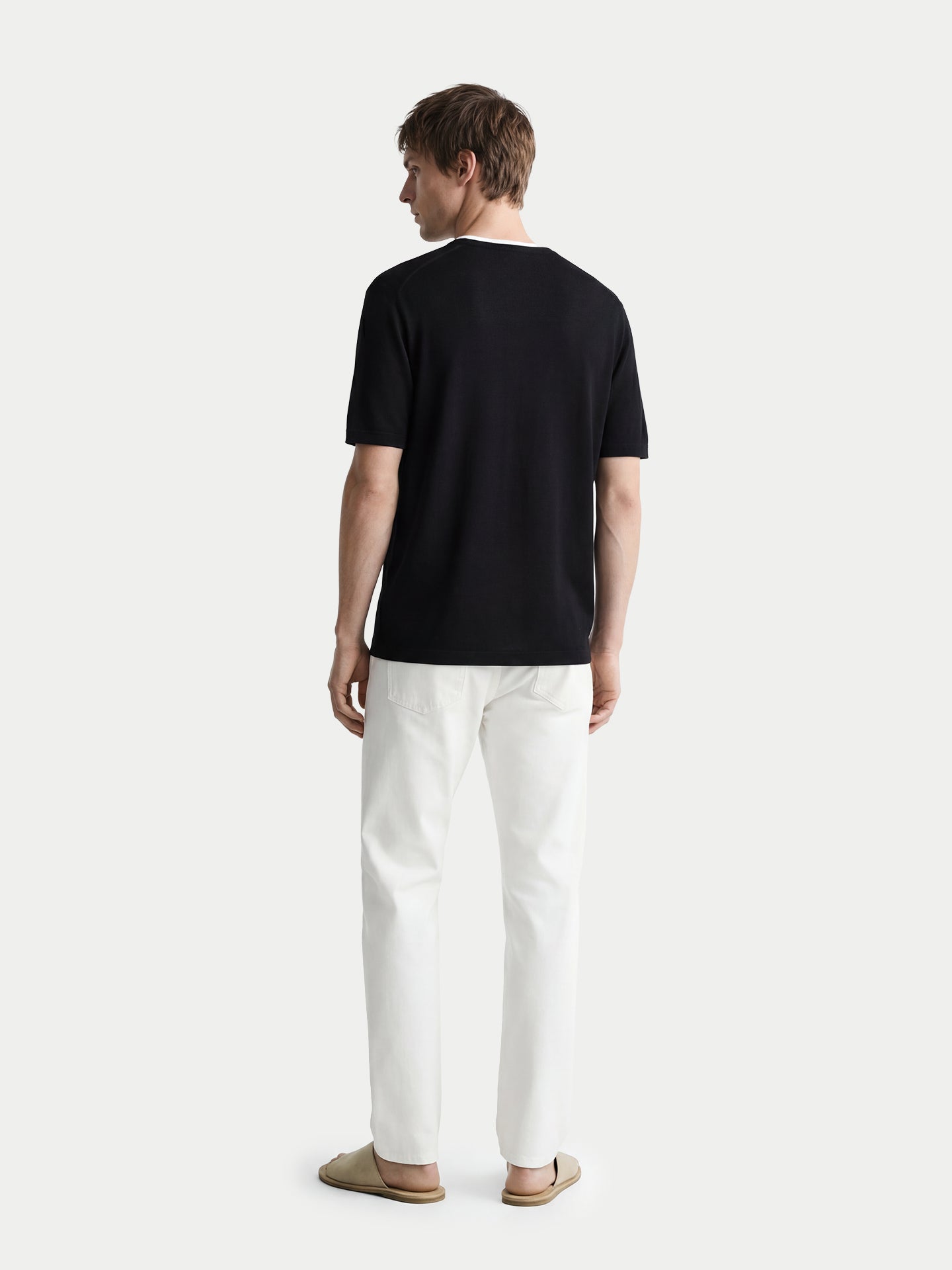 Vue de dos d'un homme portant un t-shirt en cachemire noir à col rond blanc et un pantalon blanc sur fond gris clair. #color_noir