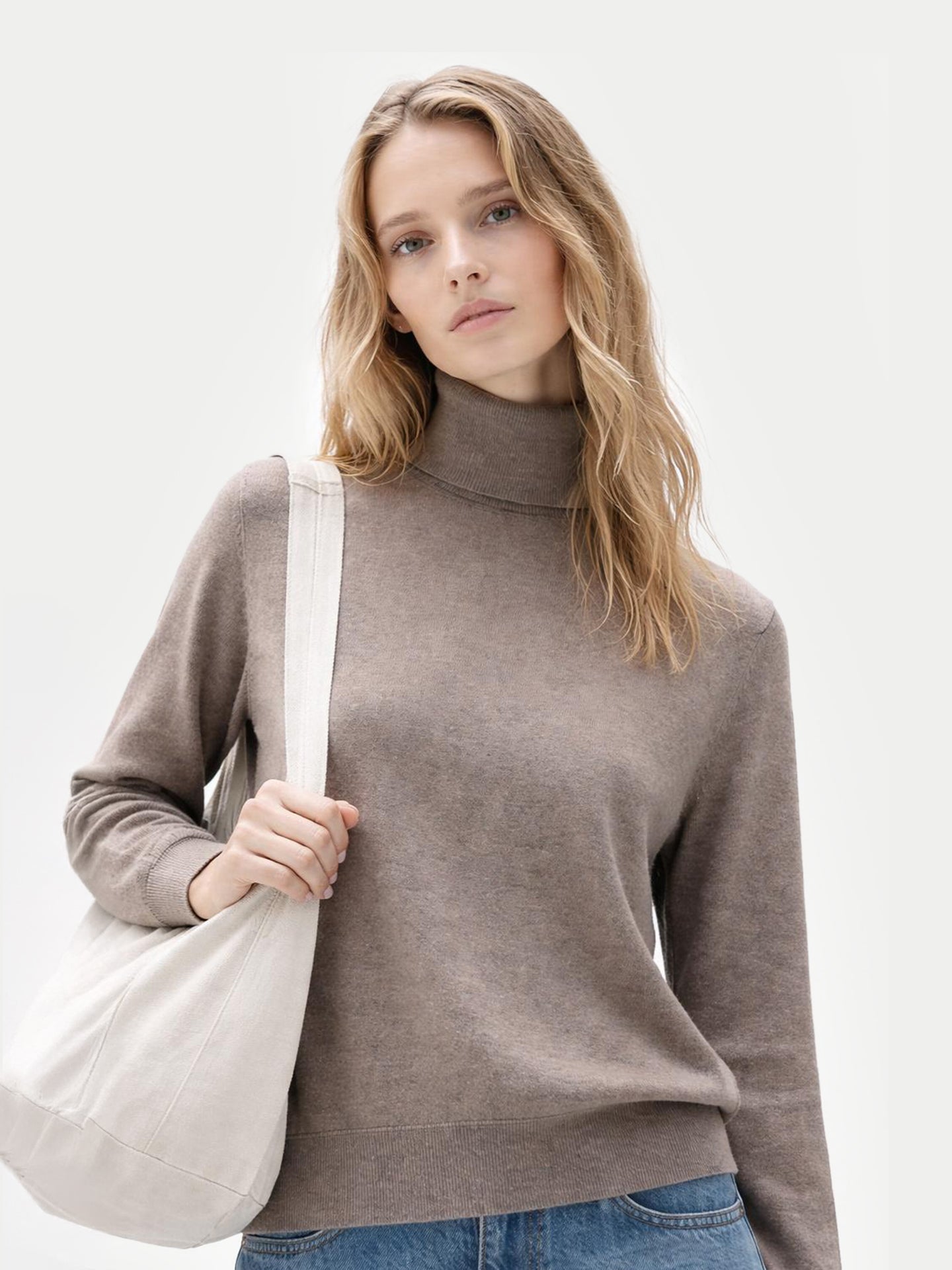 Gros plan de face d'une femme portant un pull à col roulé marron et tenant un sac cabas blanc sur un fond gris clair. #color_taupe