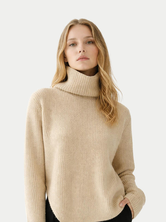 Gros plan de face d'une femme portant un pull beige côtelé à col roulé ample sur fond gris clair. #color_beige