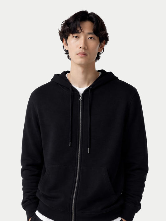 Gros plan de face d'un homme portant un sweat à capuche noir zippé avec poche latérale sur fond gris clair. #color_noir