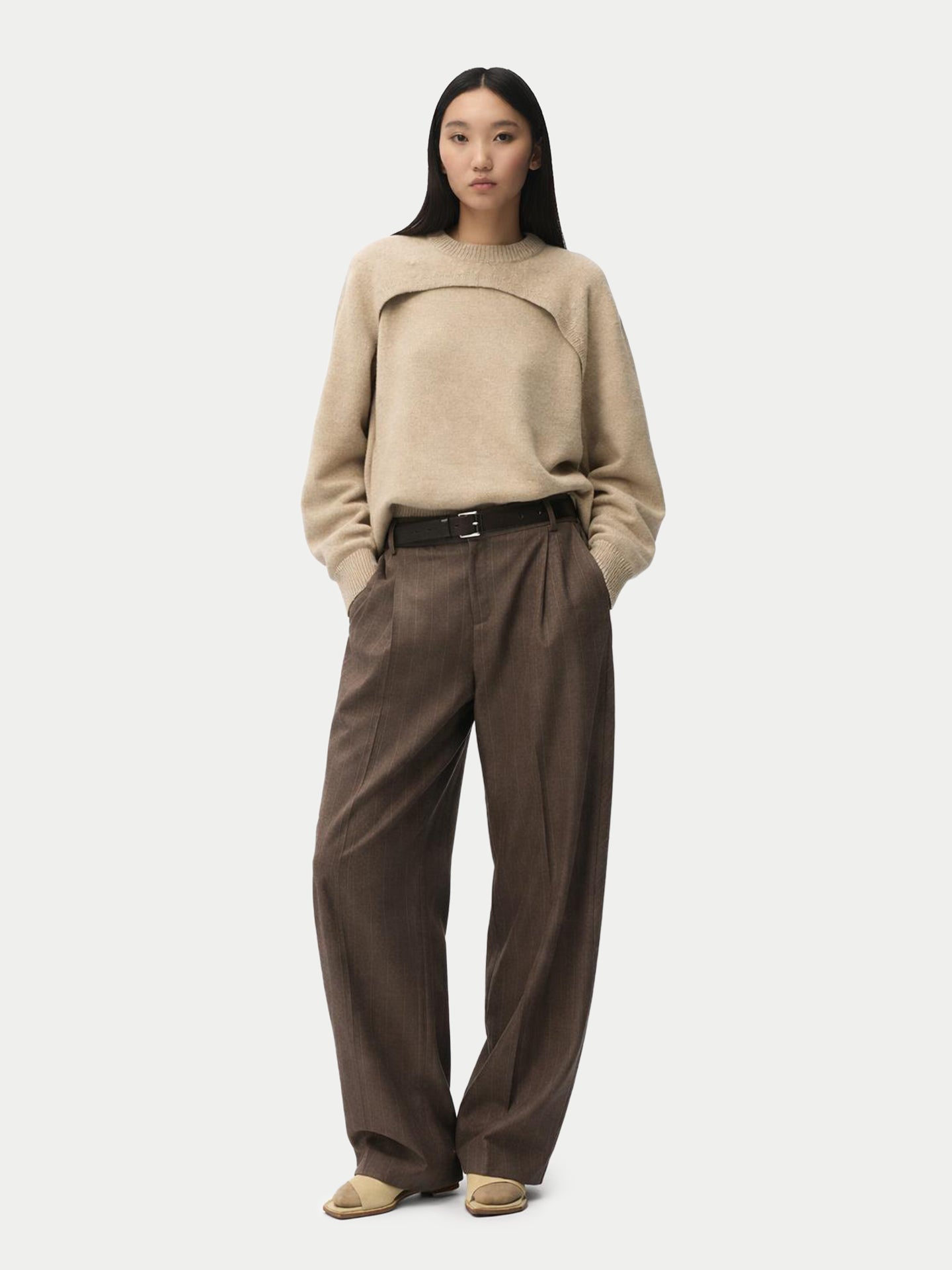 Vue de face d'une femme portant un pull beige en maille à motif architectural avec un empiècement sur la poitrine, associé à un pantalon en laine à fines rayures et une ceinture noire. #color_gris-chaud