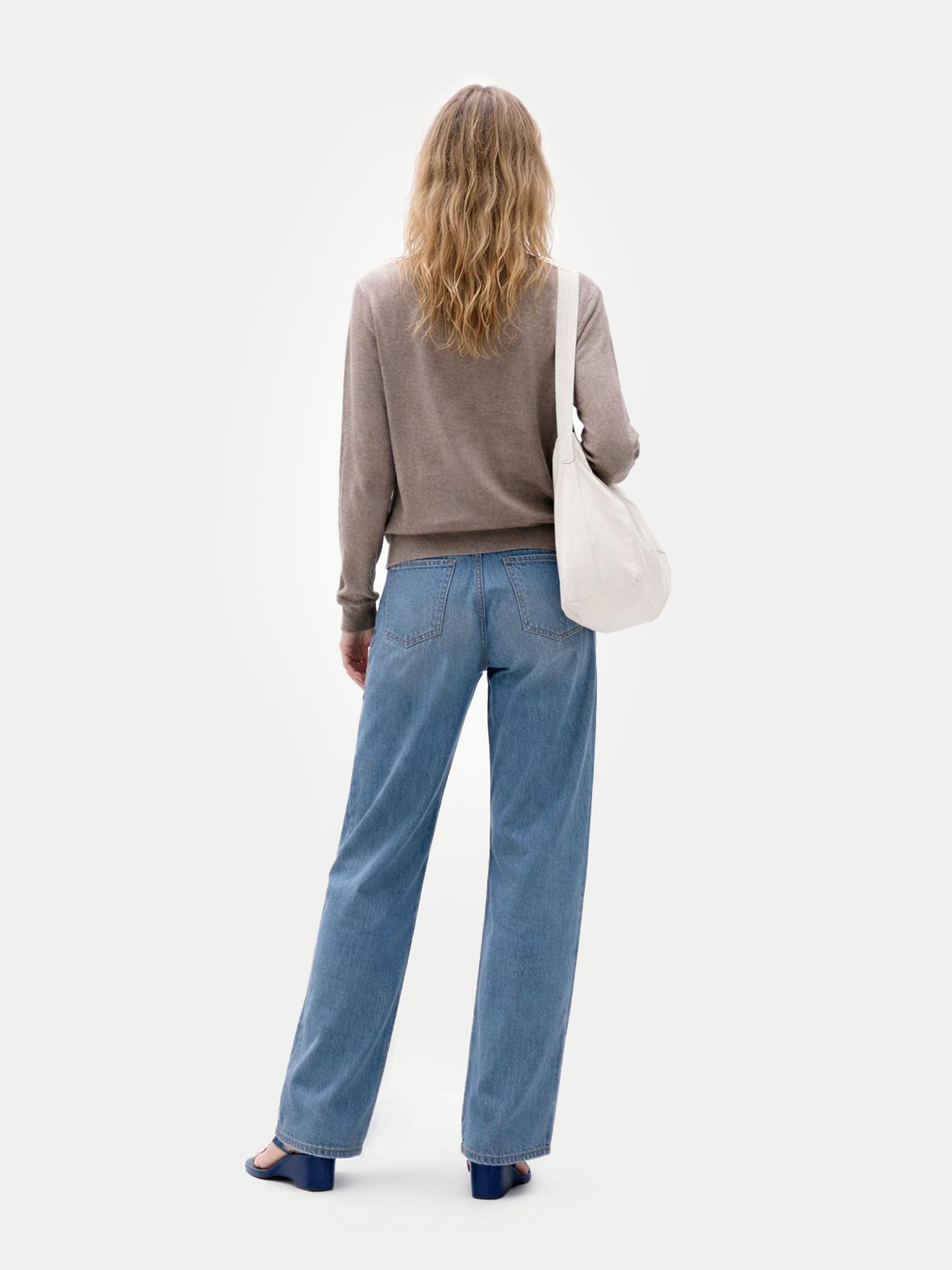 Vue de dos d'une femme portant un pull marron, un jean bleu et tenant un sac cabas blanc sur un fond gris clair. #color_taupe