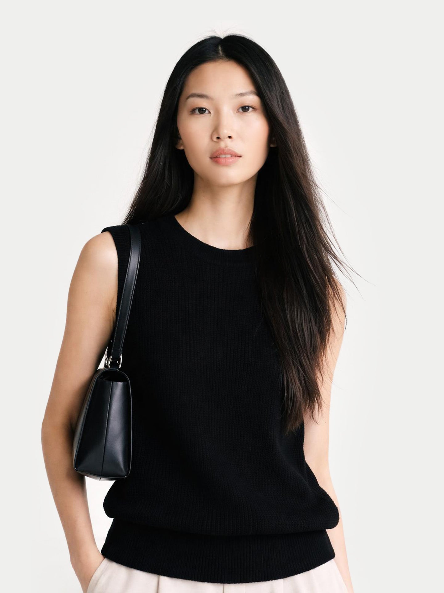 Gros plan de face d'une femme portant un débardeur noir à col rond et tenant un sac à main sur fond gris clair. #color_noir