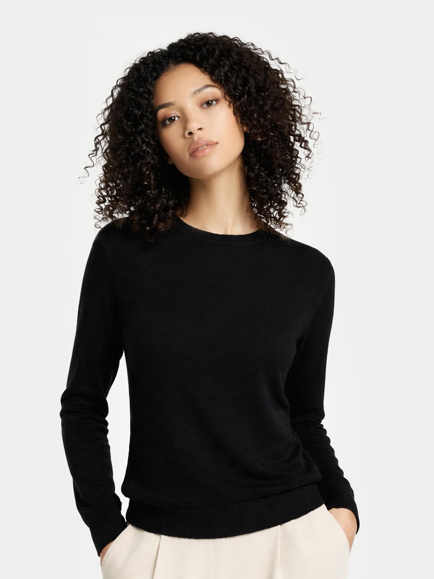 Gros plan de face d'une femme portant un pull noir à col rond et un pantalon blanc sur fond gris clair. #color_noir