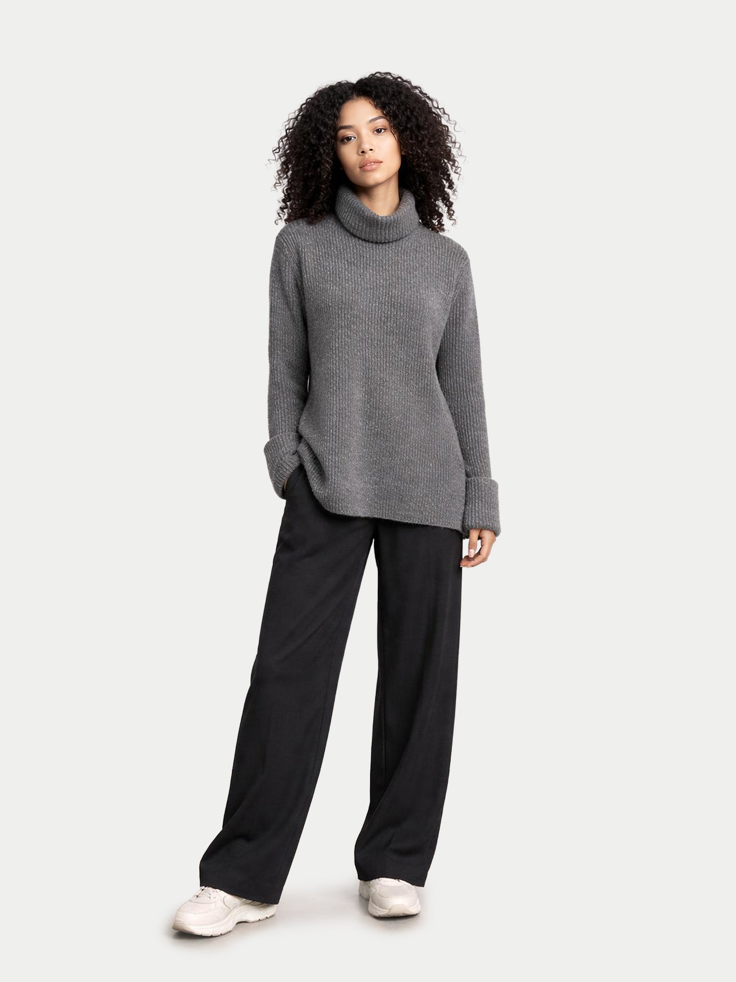 Vue de face d'une femme portant un pull gris côtelé à col roulé ample et manches retroussées, et un pantalon noir, sur fond gris clair. #color_gris-pierre