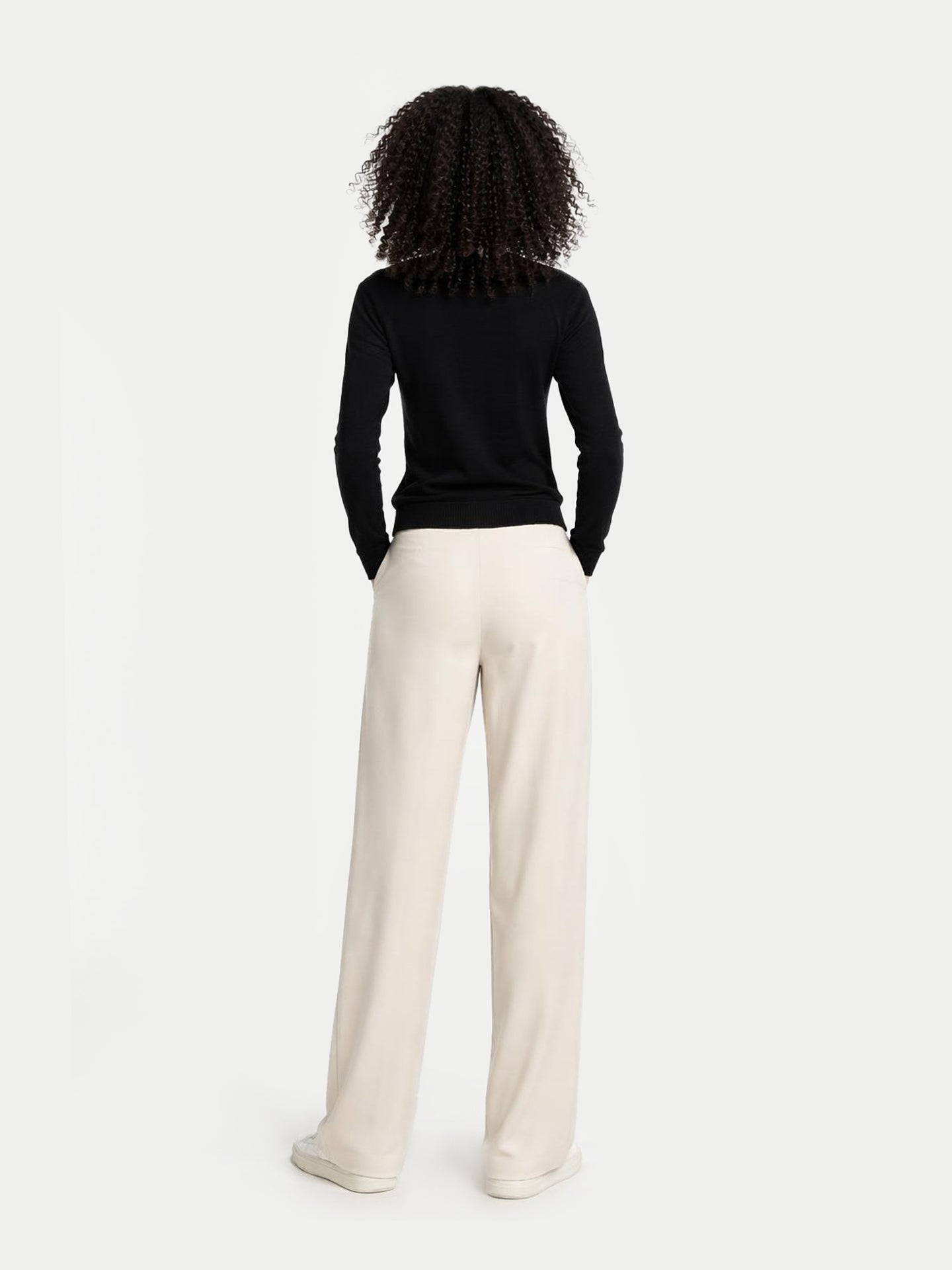 Vue de dos d'une femme portant un pull noir à col rond et un pantalon blanc sur fond gris clair. #color_noir