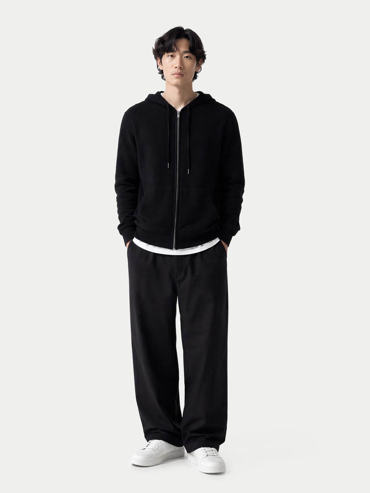 Vue de face d'un homme portant un sweat à capuche noir zippé sur un t-shirt et un pantalon blancs, sur fond gris clair. #color_noir